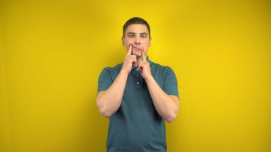 Genç bir adam yüzünde üzgün ve neşeli bir gülümseme yapar. Kadının parmakları sarı arka plandadır. Yeşil polo giyen adam..