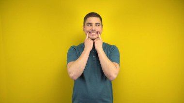 Genç bir adam, sarı arka planda parmaklarıyla neşeli bir gülümseme sergiliyor. Yeşil polo giyen adam..