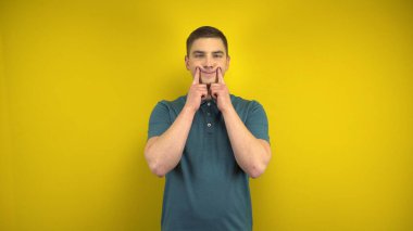 Genç bir adam, sarı arka planda parmaklarıyla neşeli bir gülümseme sergiliyor. Yeşil polo giyen adam..