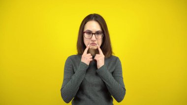 Genç bir kadın, sarı arka planda parmaklarıyla üzgün bir gülümseme sergiliyor. Gözlüklü ve yeşil kazaklı bir kadın..