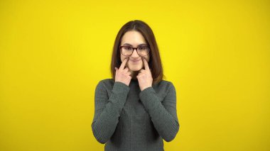 Genç bir kadın, sarı arka planda parmaklarıyla neşeli bir gülümseme sergiliyor. Gözlüklü ve yeşil kazaklı bir kadın..