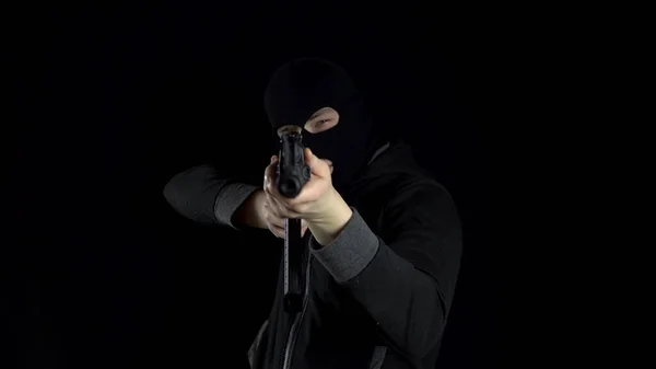 A man in a balaclava mask stands with an AK-47 assault rifle. The bandit stands with a gun on his shoulder, and then aims at the camera. On a black background.