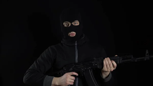 A man in a balaclava mask stands with an AK-47 assault rifle. The bandit charges the machine and stands. On a black background.