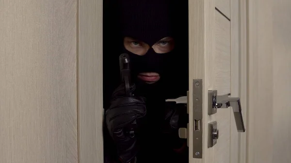 A robber is standing behind the door with a pistol close-up. The masked thug slowly opened the door and looks at the camera.