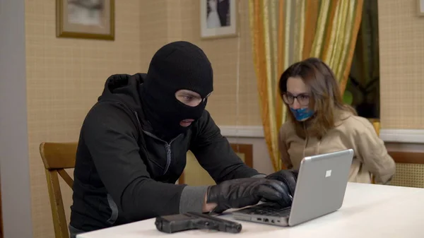 A burglar is trying to break into a laptop by taking a young woman hostage. A masked thug sits in a house and cannot break into a laptop and threatens a hostage with a gun. Theft of data from a