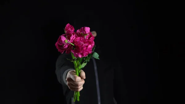 A man in a balaclava mask is standing with flowers. The bandit holds out a bouquet of pink flowers to the camera. On a black background.