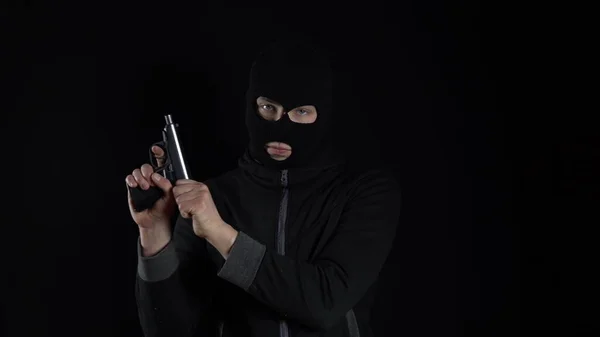A man in a balaclava mask stands with a gun. The bandit reloaded the gun and stands on a black background.