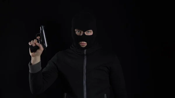 A man in a balaclava mask stands with a gun. The bandit reloaded the gun and stands on a black background.