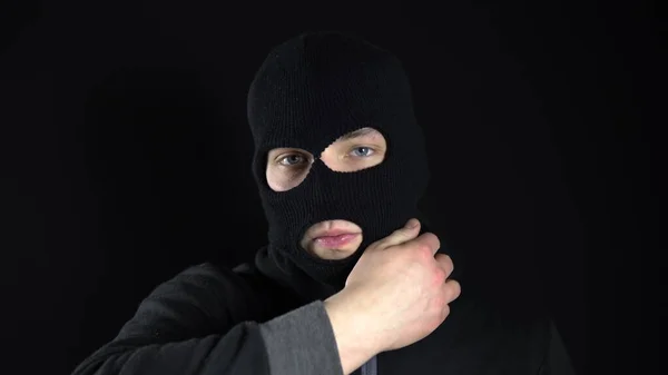 A young man takes off the mask of a balaclava. Bandit on a black background close-up.