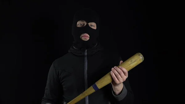 A man in a balaclava mask is standing with a baseball bat. A gangster is holding a baseball bat on a black background.