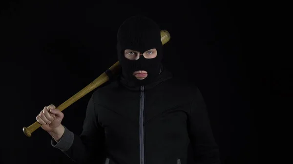 A man in a balaclava mask is standing with a baseball bat. A bandit stands on a black background with a baseball bat over his shoulder.