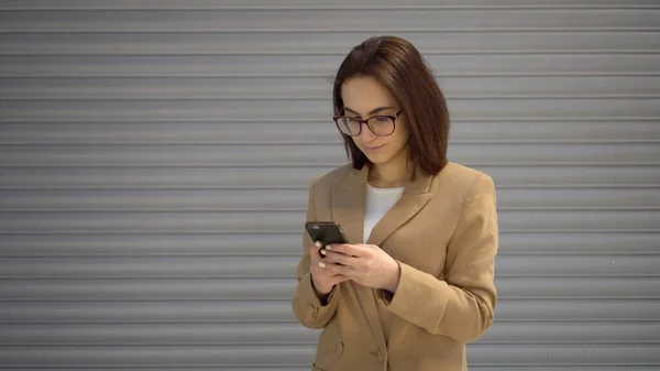 A young woman stands on a gray background and writes on the phone.