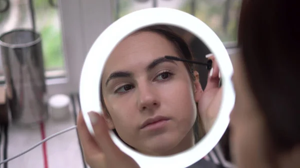 A young woman looks into a small illuminated mirror and paints her eyelashes with mascara. View of the girl through the mirror. Stock Picture