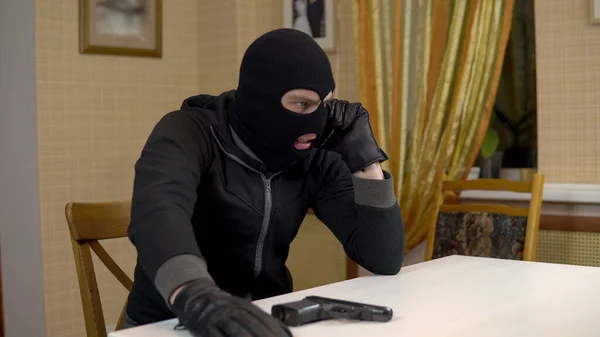 Burglar talking on the phone. A masked thug sits in a house and threatens the telephone with a gun. Extortion by phone. Royalty Free Stock Images