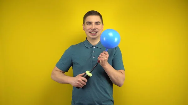 A young man inflates a blue balloon with a pump on a yellow background. Man in a green polo. Stock Photo