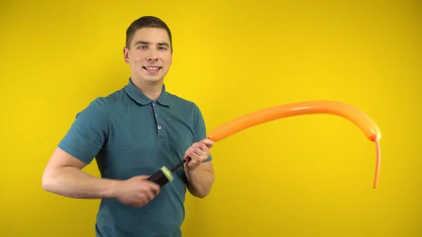 A young man inflates a long orange balloon with a pump on a yellow background. Man in a green polo. Stock Picture