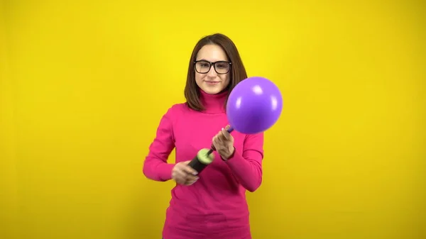 一位年轻女子用黄色背景的水泵充气紫色气球。穿着粉色套头毛衣和眼镜的女孩. 免版税图库图片