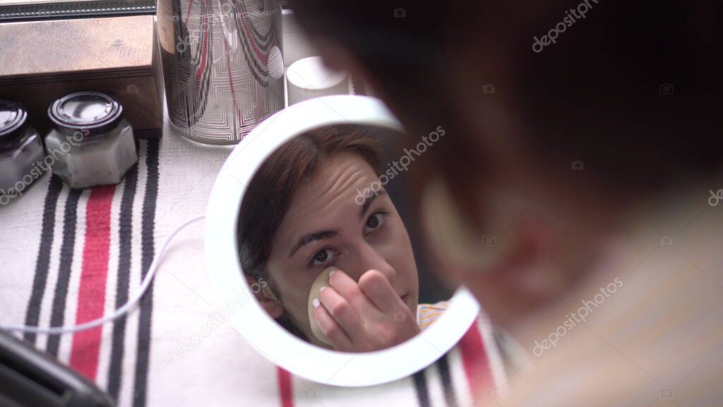 A young woman in front of a window looks into a small mirror with light and applies makeup with a sponge. View of the girl through the mirror.