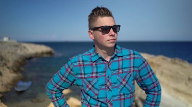 Genç bir adam denizin arka planında duruyor. Mavi gömlekli bir adam güneş gözlüğü takıyor. Etraftaki doğayı keşfeder