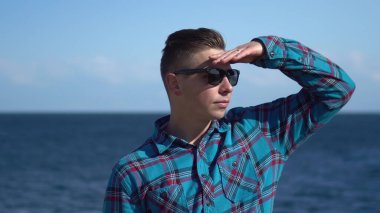 Denizin arka planında genç bir adam. Güneşli havada güneş gözlüklü bir adam. Etraftaki doğayı keşfeder