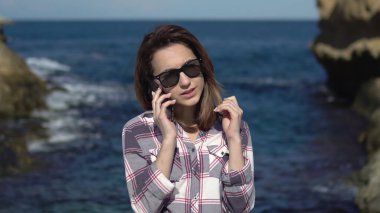 Genç bir kadın denizin arka planındaki akıllı telefondan arama yapıyor. Tatilde telefonu olan bir kadın. Güneşli havada mavi deniz