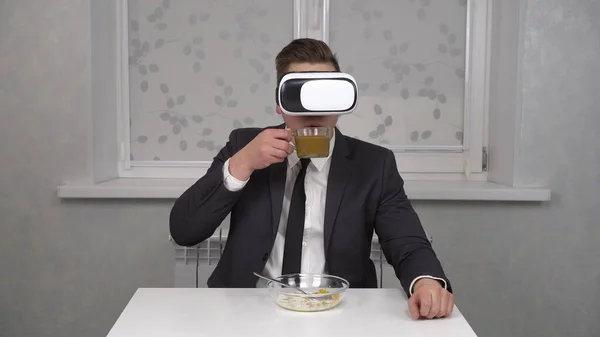 A young man in a suit and a VR helmet eats cereal and drinks coffee. Young businessman at home in the morning kitchen.