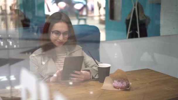 En ung kvinna sitter bakom ett glas på ett kafé med en tablett i händerna. Flickan dricker kaffe och arbetar. — Stockvideo