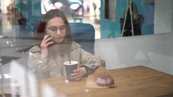 En ung kvinna bakom glaset sitter på ett kafé och pratar i telefon. Flickan dricker kaffe.. — Stockvideo