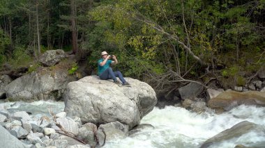 Panama şapkalı genç bir adam bir taşın üzerine oturur ve nehir kenarındaki dağlarda telefonuyla fotoğraf çeker..