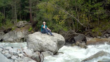 Panama şapkalı genç bir adam bir taşın üzerinde oturur ve nehir kenarındaki dağlarda telefonla konuşur. Dağlara git..