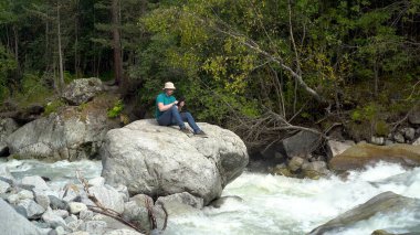 Panama şapkalı genç bir adam nehir kıyısındaki dağlarda elinde tabletle bir taşın üzerinde oturuyor. Dağlara git..