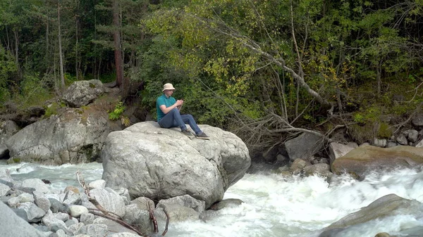 Młody człowiek w kapeluszu Panamy siedzi na kamieniu z telefonem w rękach w górach nad rzeką.. — Zdjęcie stockowe