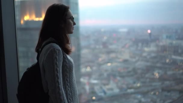 Mujer asiática joven con gafas mira por la ventana con un rascacielos en la noche. Chica en un apartamento con ventanas panorámicas. — Vídeos de Stock