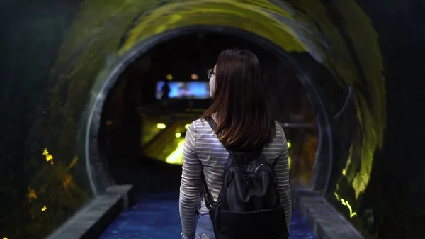 Uma jovem mulher com óculos caminha pelo túnel no oceanário. A menina olha para a vida marinha por trás do vidro. — Fotografia de Stock