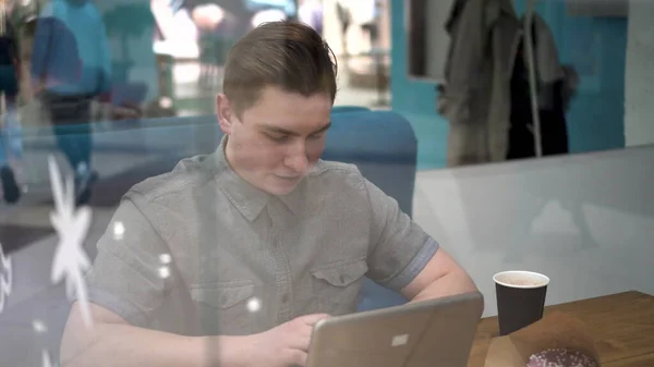 Jeune homme assis derrière le verre dans un café avec un ordinateur portable. L'homme boit du café et travaille. — Photo