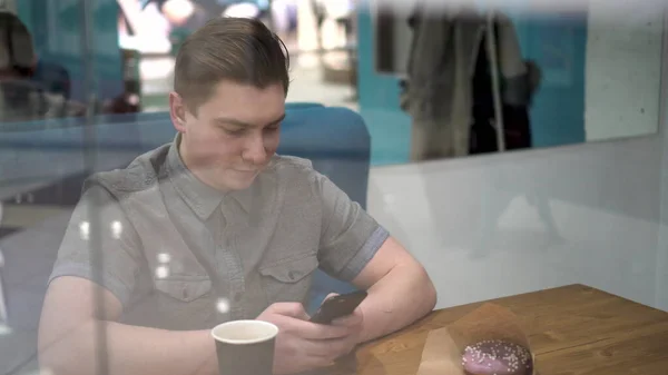 Un giovane uomo è seduto dietro un bicchiere in un caffè con un telefono in mano. L'uomo beve caffè e lavora. — Foto Stock