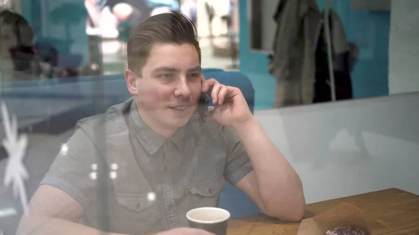 Um jovem atrás do vidro senta-se em um café e fala ao telefone. O homem está a beber café.. — Fotografia de Stock