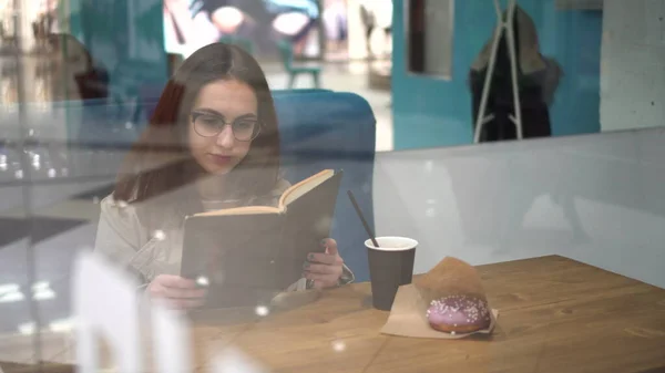 Mladá žena sedí za sklenicí v kavárně a čte si knihu. Ta holka pije kafe.. — Stock fotografie