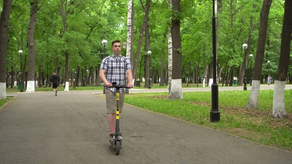 Un joven europeo con camisa y pantalones cortos monta un scooter eléctrico en el parque. —  Fotos de Stock