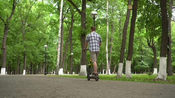 Un joven europeo con camisa y pantalones cortos monta un scooter eléctrico en el parque. Vista inferior. —  Fotos de Stock