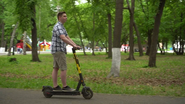 Egy fiatal európai férfi ingben és rövidnadrágban elektromos robogóval a parkban.. — Stock Fotó