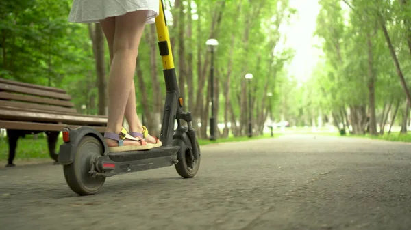 Jeune femme en robe blanche monte un scooter électrique de partage dans le parc. Vue du bas. — Photo