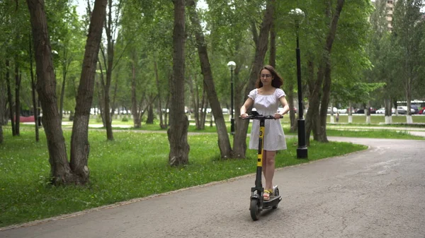 Jonge Aziatische vrouw in een witte jurk rijdt een delende electro scooter in het park. Meisje met bril. — Stockfoto