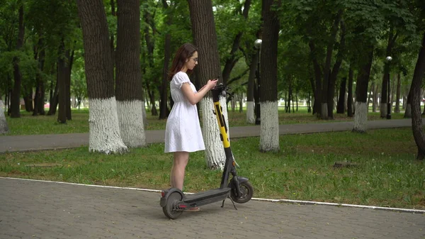 Ung kvinna i vit klänning börjar en åktur på en delad elskoter i parken. Flicka i glasögon. — Stockfoto