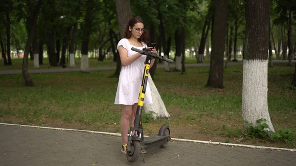 Mujer asiática joven en un vestido blanco comienza un paseo en un scooter electro compartido en el parque. Chica en gafas. —  Fotos de Stock