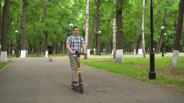 Un joven europeo con camisa y pantalones cortos monta un scooter eléctrico en el parque. — Vídeo de stock