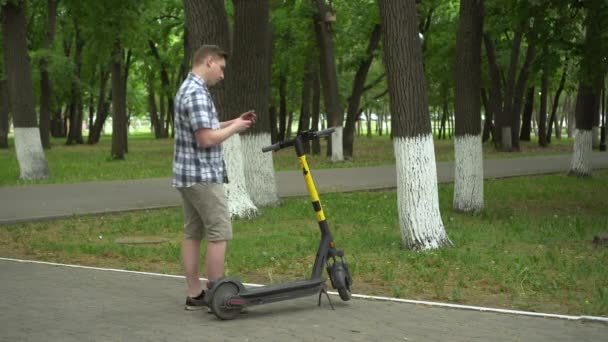 Een jonge Europeaan in overhemd en korte broek begint een ritje op een elektrische scooter in het park. — Stockvideo