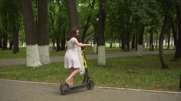 Uma jovem mulher em um vestido branco completa um passeio em uma partilha de e-scooter no parque. Menina de óculos. — Vídeo de Stock