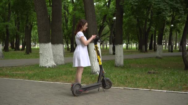 Eine junge Frau im weißen Kleid beginnt eine Fahrt auf einem Elektro-Roller im Park. Mädchen mit Brille. — Stockvideo