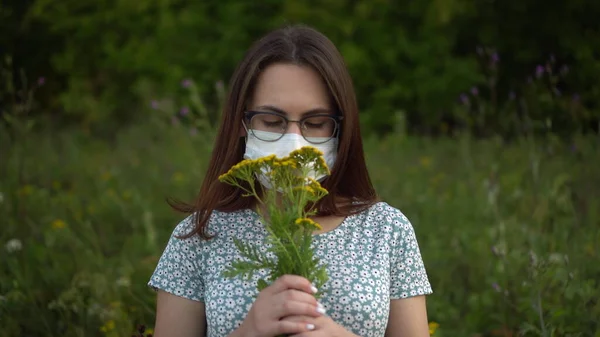 Mladá žena v lékařské masce očichává květiny a potřásá hlavou, protože nesmrdí. Dívka v brýlích a šaty v přírodě. — Stock fotografie